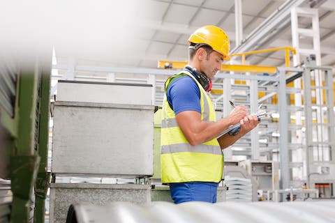 Manufacturing worker going over record keeping while in the factory.  The worker is a white male wearing a yellow safety jacket with a yellow hard hat. He is holding a clipboard in his left hand and has a pen in  his right hand. He had a blue short sleeve shirt on and safety earphones around his neck.  image